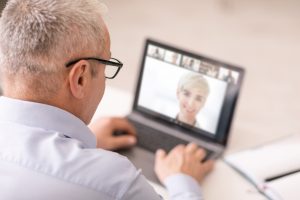 Distant Virtual Meeting Concept. Back over the shoulder view of senior mature man sitting at desk, looking at pc screen at home office, making online video call, talking on web, selective focus